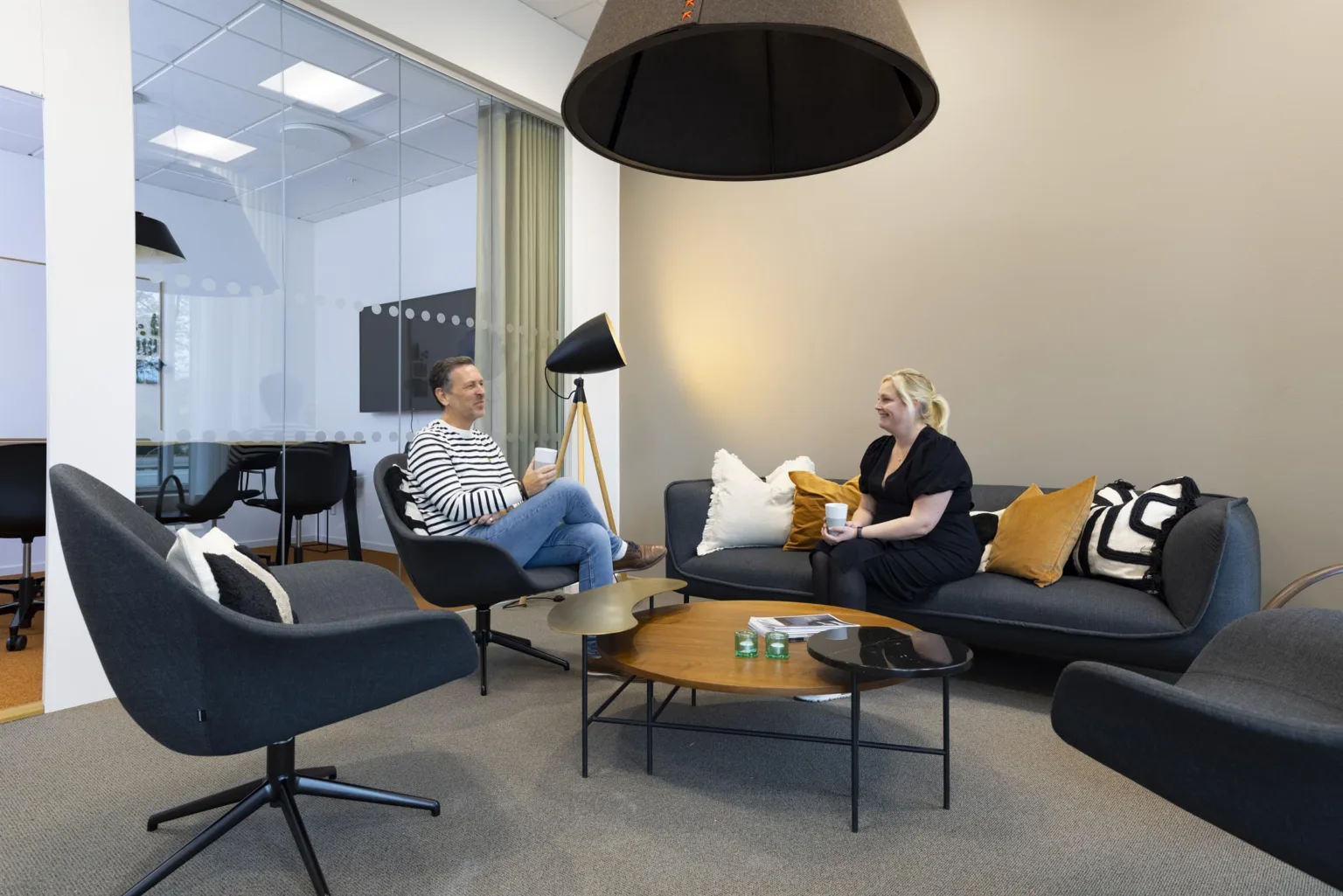 3. Två personer sitter och samtalar i en lounge med soffor, kuddar och en taklampa.