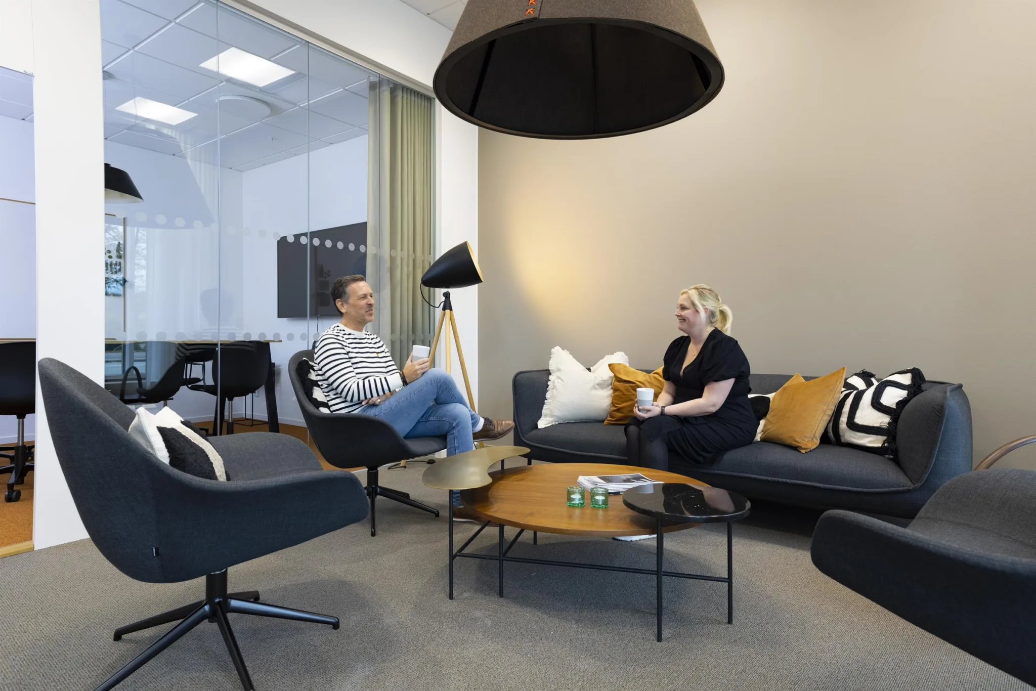 3. Två personer sitter och samtalar i en lounge med soffor, kuddar och en taklampa.