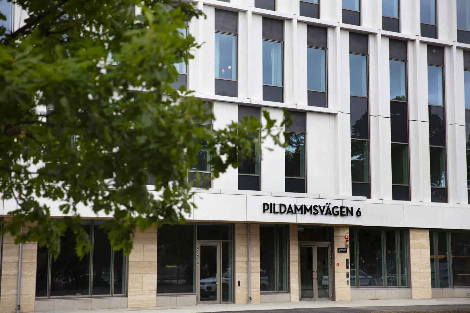 1. En modern byggnad med vit fasad och texten “Pildammsvägen 6” ovanför entrén, omgiven av grönska.