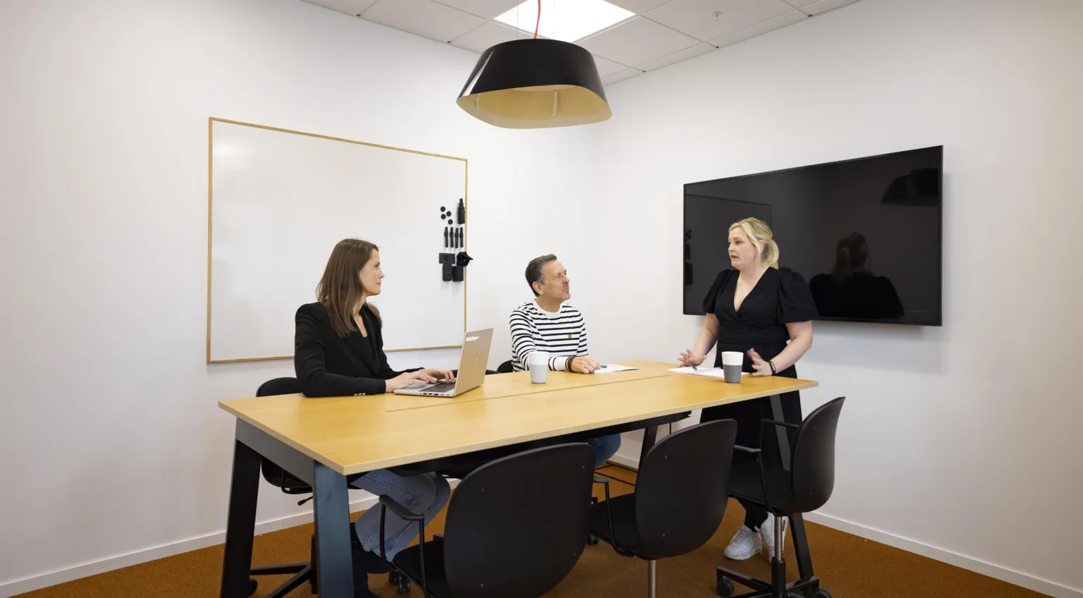 5. Tre personer sitter runt ett konferensbord i ett mötesrum med en whiteboard och en väggmonterad skärm.