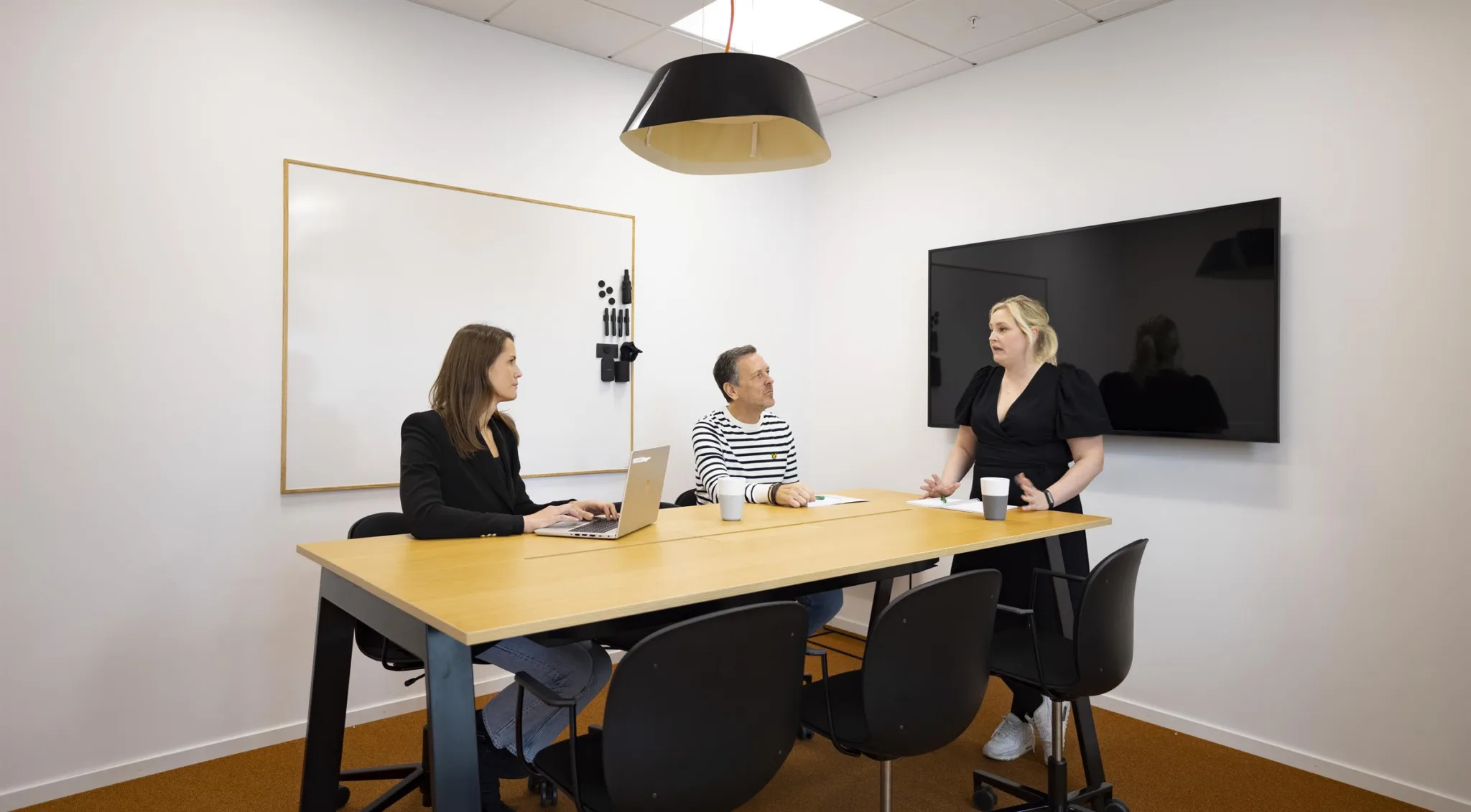 5. Tre personer sitter runt ett konferensbord i ett mötesrum med en whiteboard och en väggmonterad skärm.