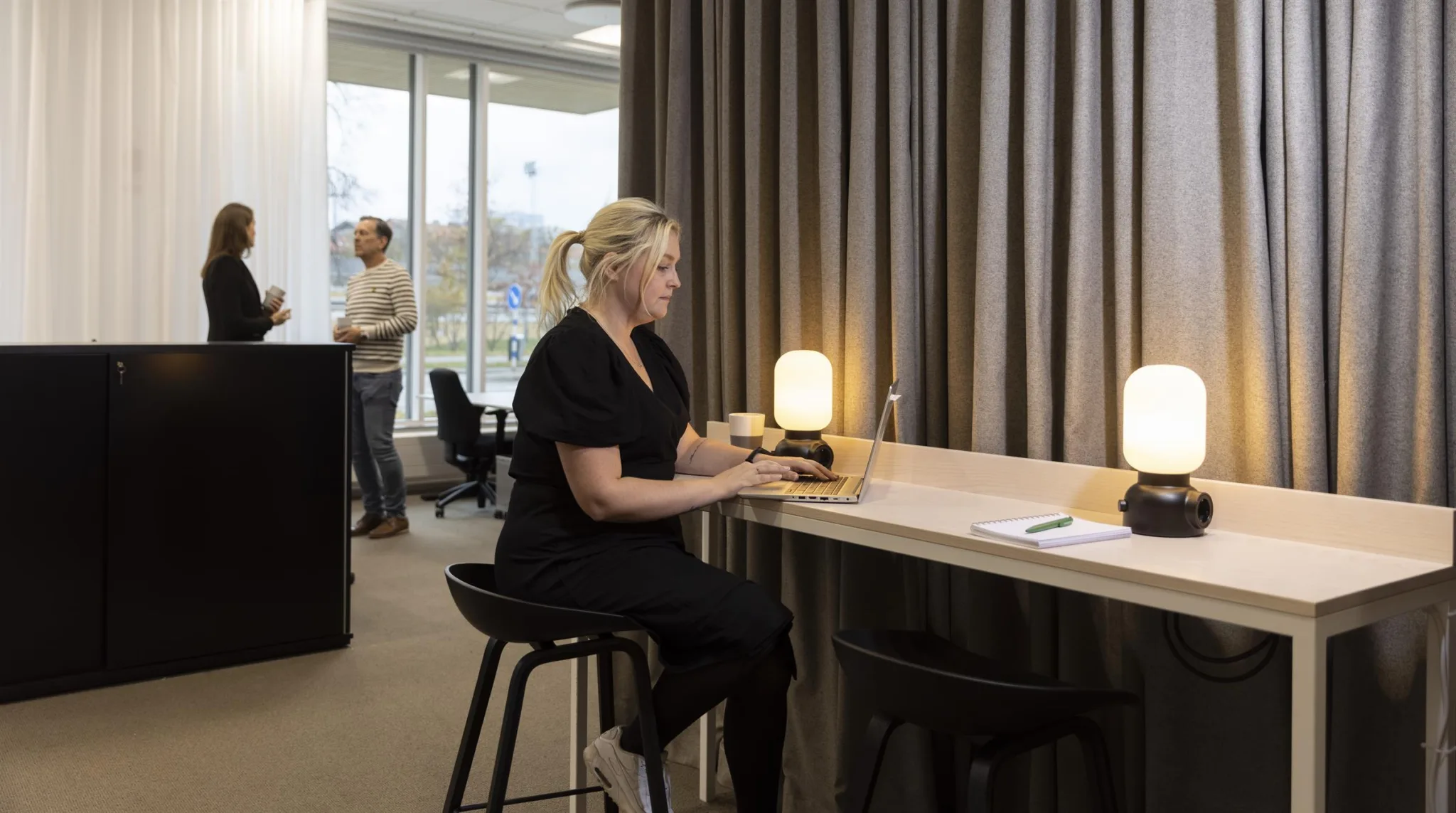 2. En kvinna sitter vid en hög arbetsbänk och arbetar på en laptop medan två personer samtalar i bakgrunden i ett kontor med stora fönster.