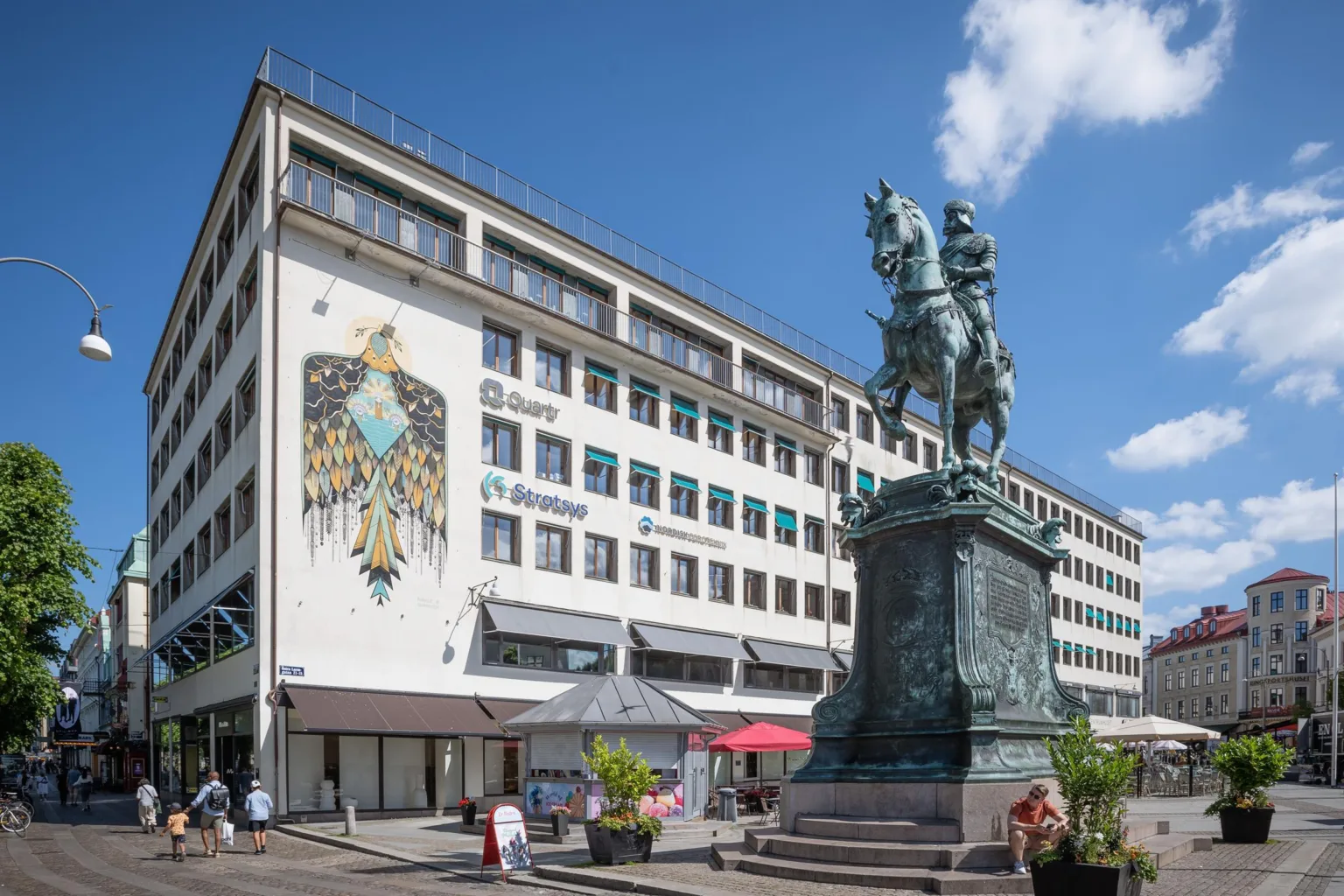 En byggnad med fasadmålning av en fågel och en ryttarstaty på ett torg under klar himmel.