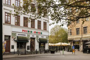 2. Fasaden av en restaurang med namnet "Piccolo Mondo", omgiven av träd och utomhusbord under parasoller.