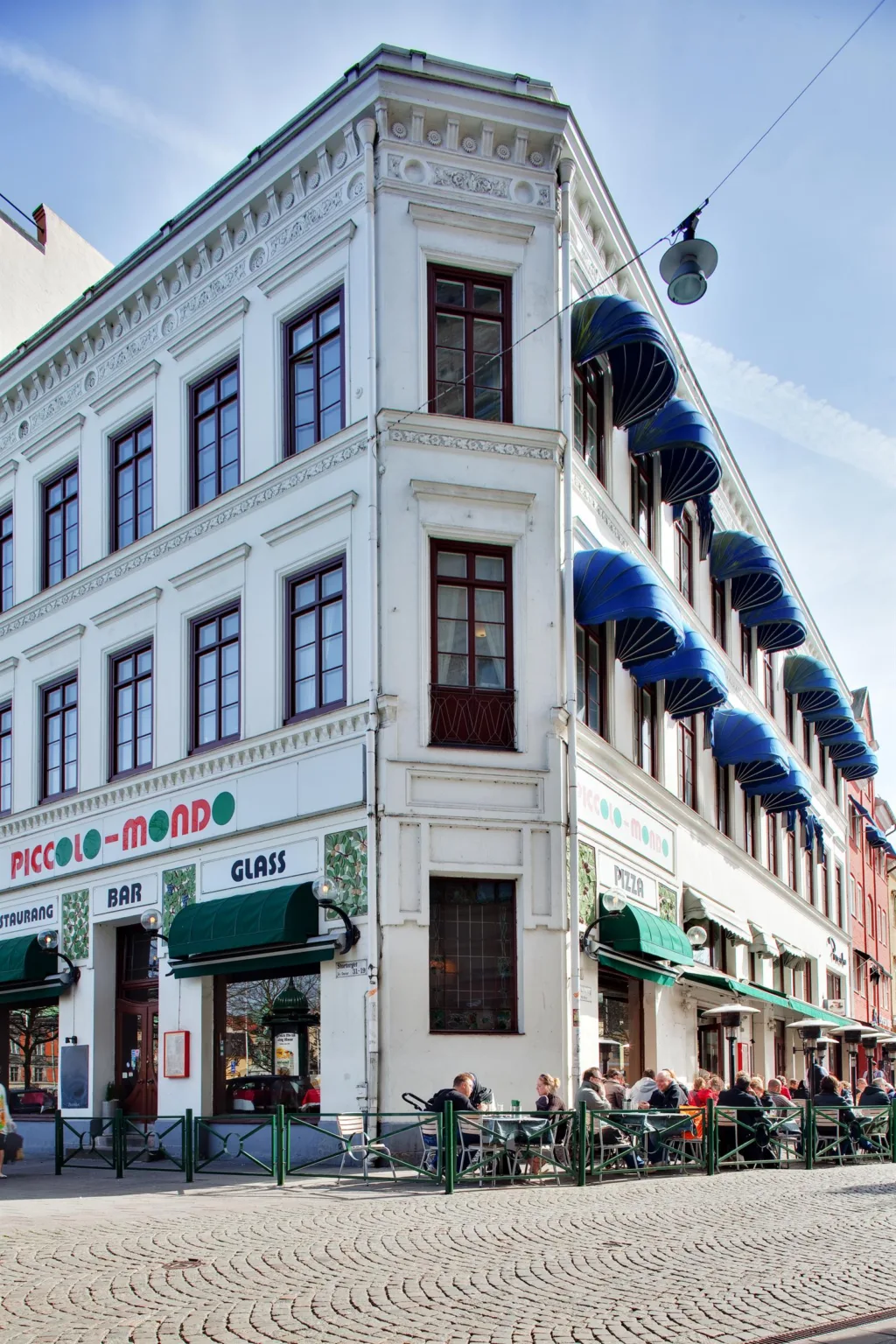 4. En vit hörnbyggnad med blå markiser och en butiksskylt för "Piccolo Mondo" samt uteservering med gröna detaljer.