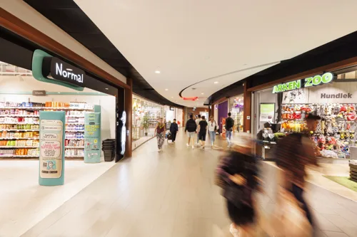 En bred gång i en shoppinggalleria med butiker på båda sidor och suddiga människor som går förbi.