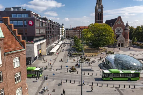 5. Ett stadslandskap med en öppet torg, gröna bussar och en kyrka i bakgrunden.