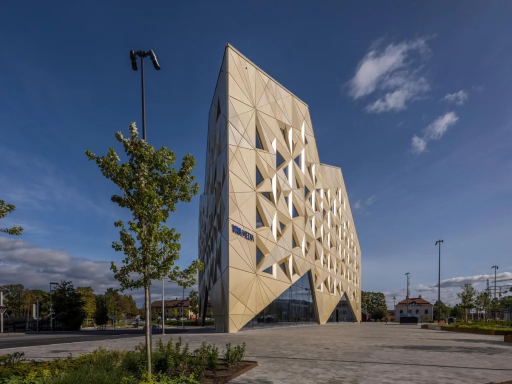 En modern byggnad med geometriska mönster på fasaden, omgiven av ett öppet område med träd och bänkar.