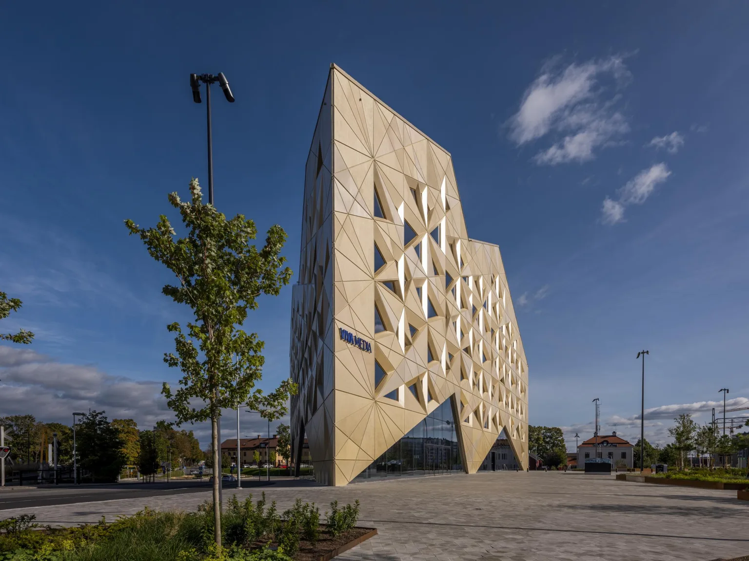 En modern byggnad med geometriska mönster på fasaden, omgiven av ett öppet område med träd och bänkar.