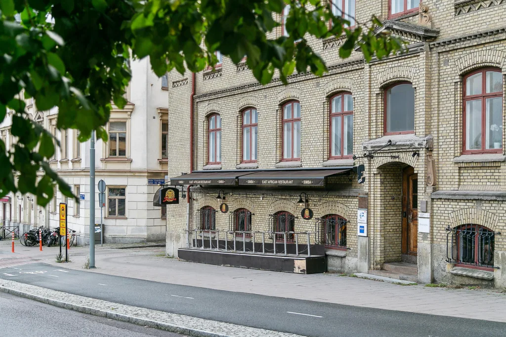 En gul tegelbyggnad med en uteservering under trädens skugga, trottoaren och gatan syns i förgrunden.