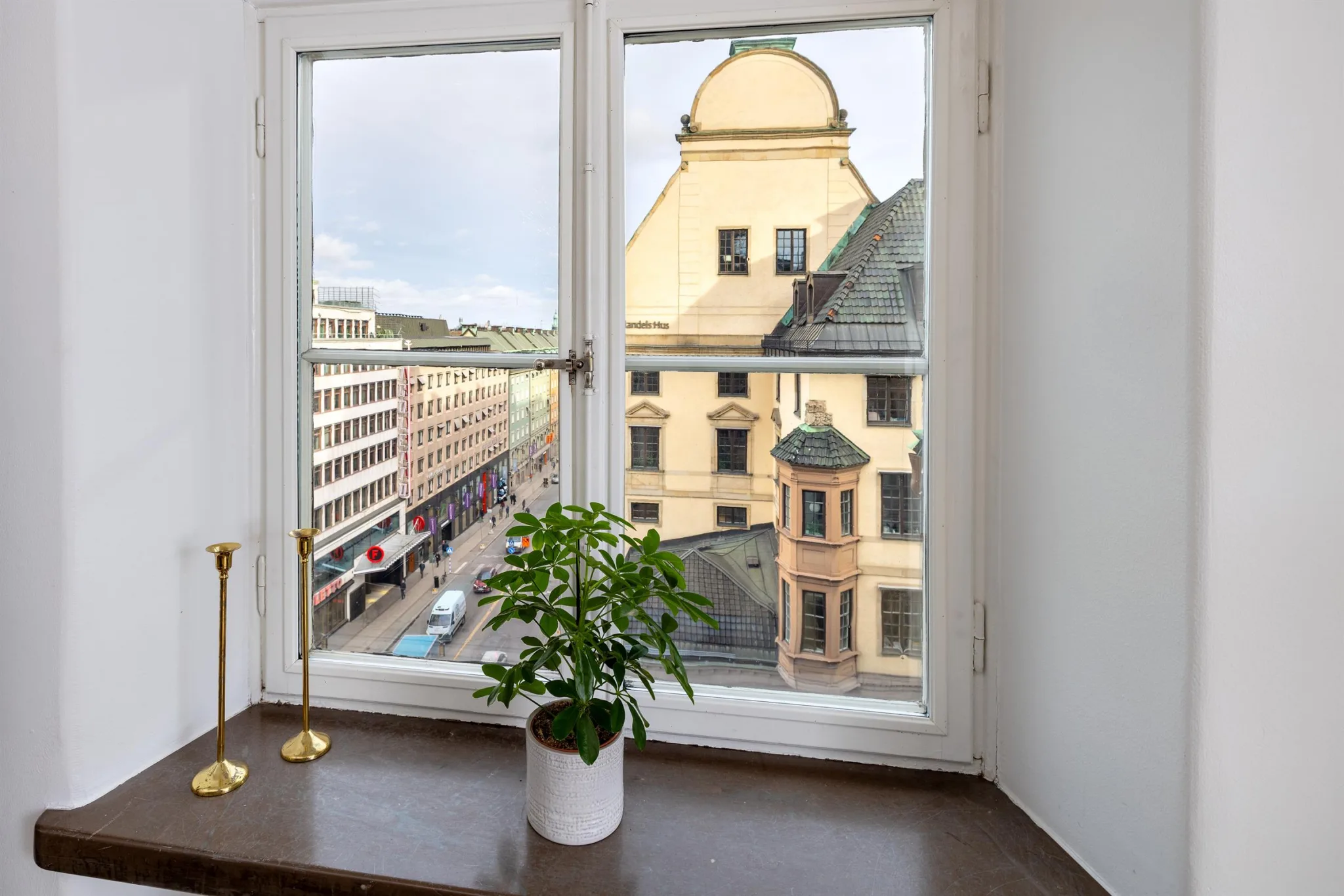 Ett fönster med utsikt över en stadsgata och en gul byggnad; en grön växt och två ljusstakar står på fönsterbrädan.