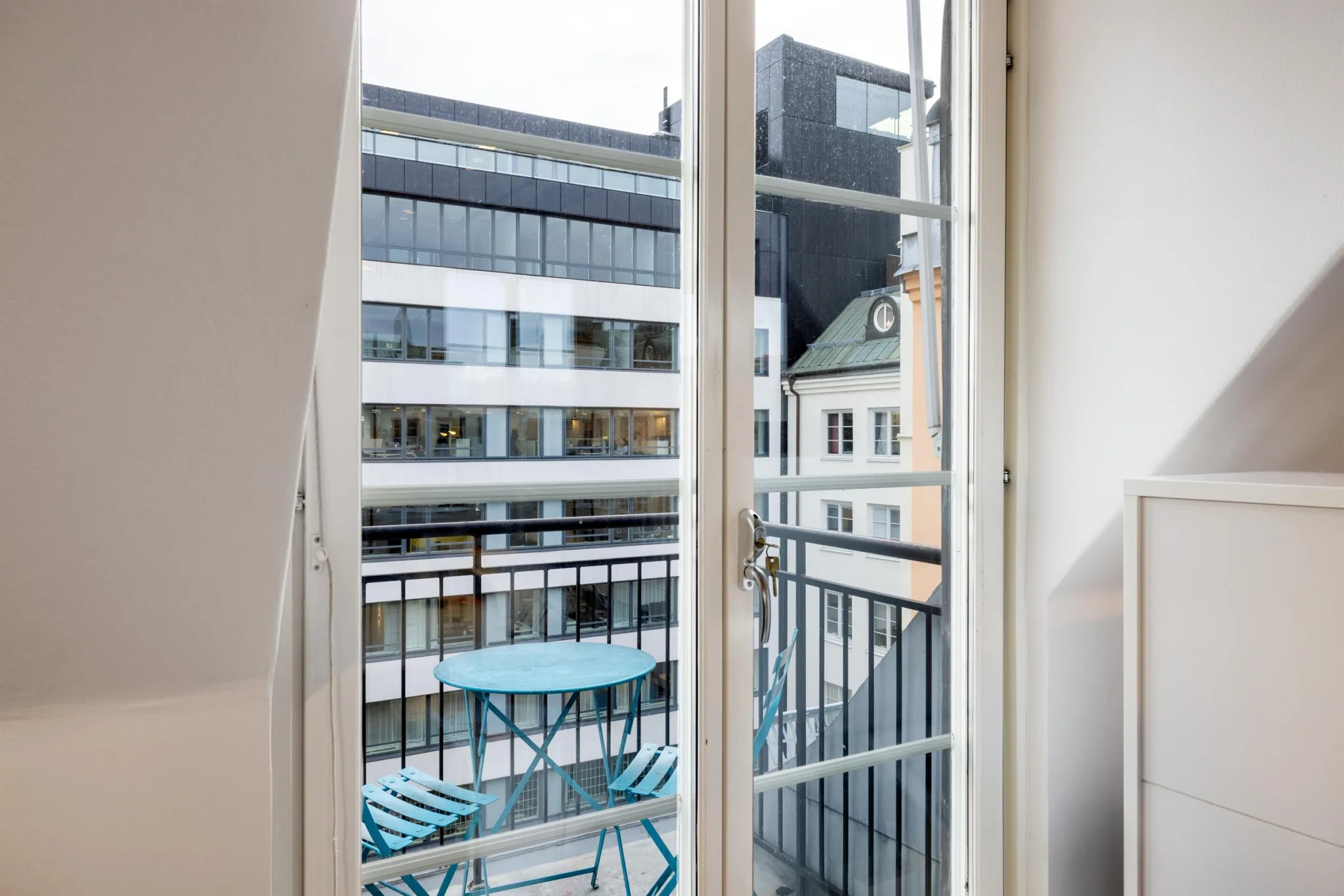 En balkong med blå bord och stolar, sett genom glasdörrar, med utsikt över moderna och traditionella byggnader.