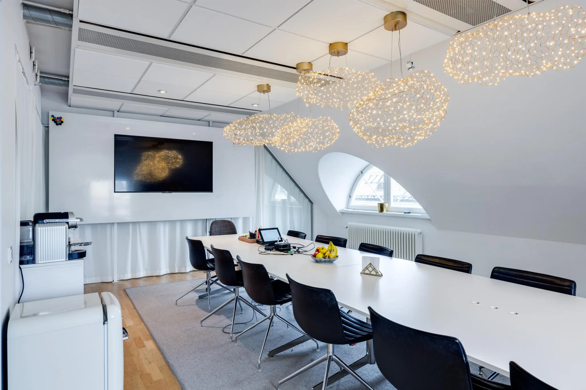 Ett konferensrum med lång vit bord, svarta stolar, dekorativa hänglampor och en väggmonterad TV.