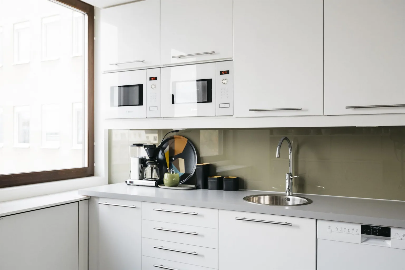 Ett modernt kök med vita skåp, en diskho, två mikrovågsugnar och en kaffemaskin.