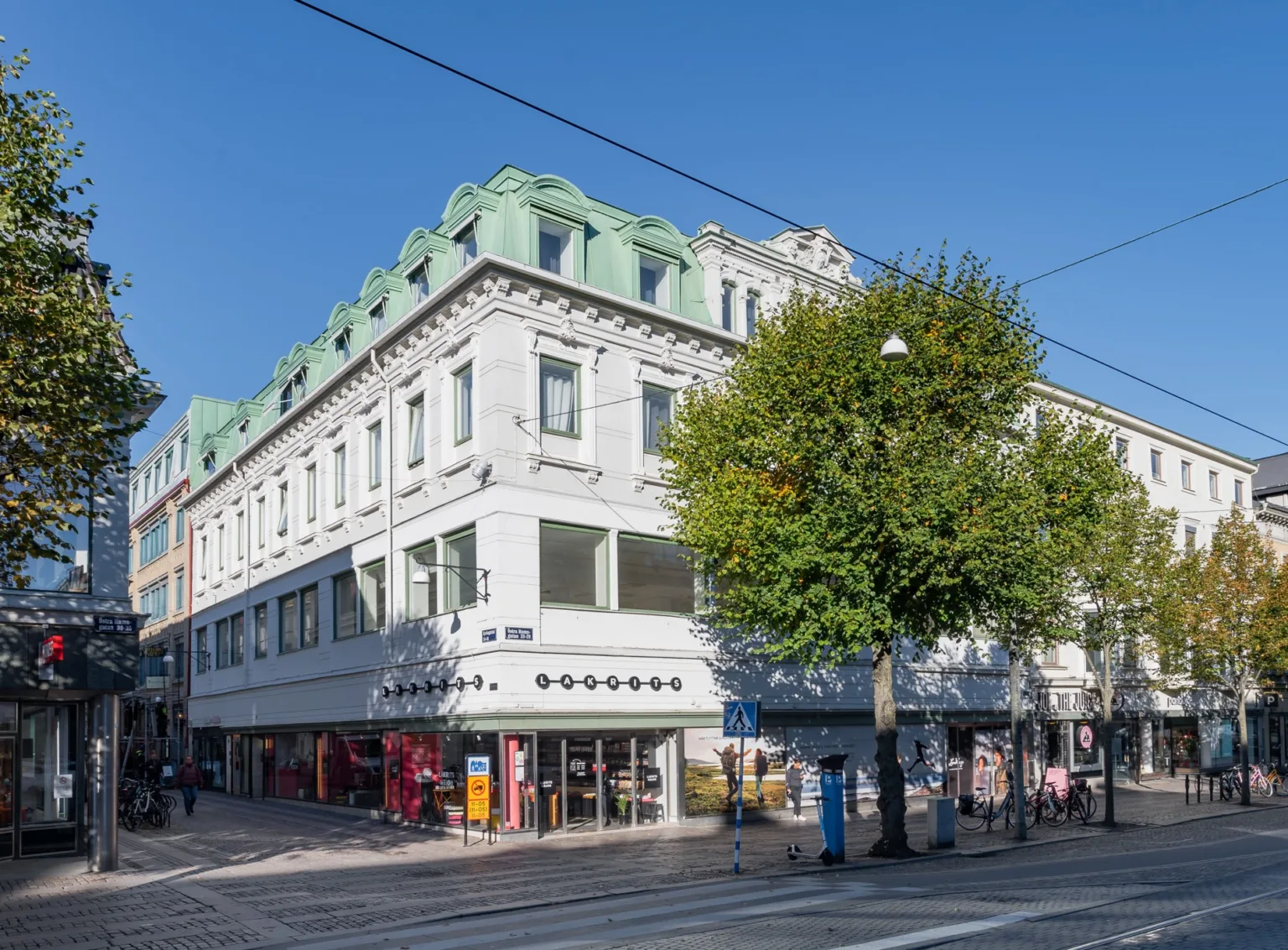 Vit byggnad med grönt tak och flera butiker på bottenvåningen, omgiven av en bred gata med ett träd.