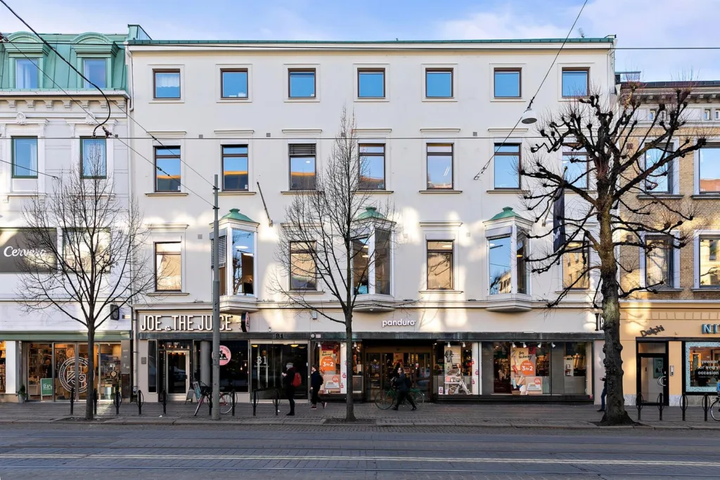 Vit fasad med fönster och butiker på bottenvåningen, träd med nakna grenar framför byggnaden.