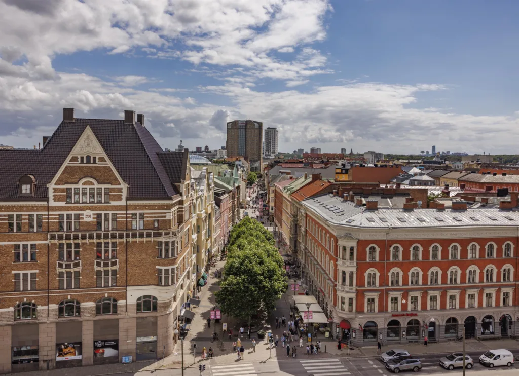 1. Vy över en stadsgata kantad av gamla tegelbyggnader med grönskande träd i mitten och höghus i bakgrunden under en delvis molnig himmel.