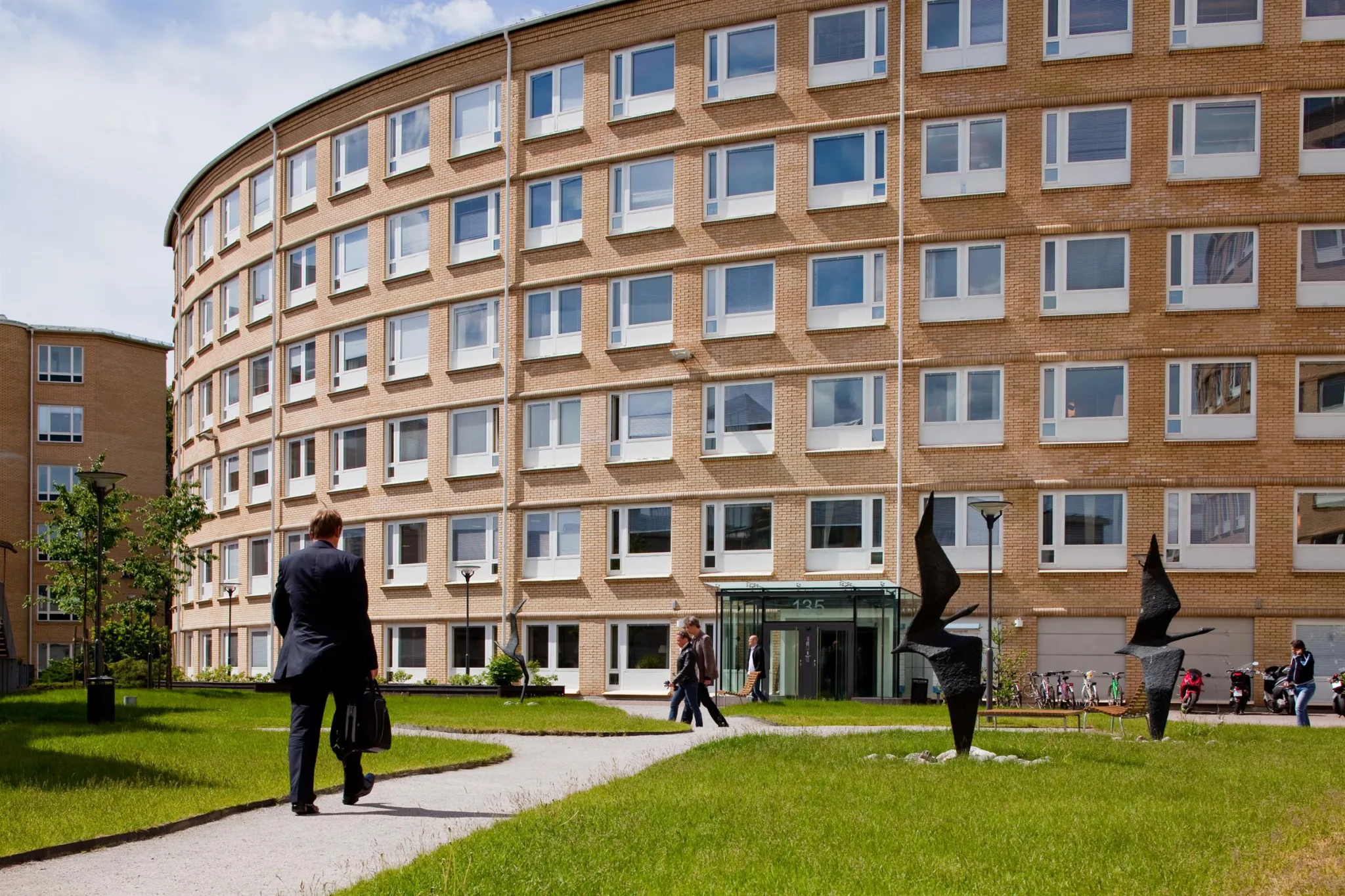 En böjd tegelbyggnad med många fönster, en gräsmatta framför och människor som promenerar.