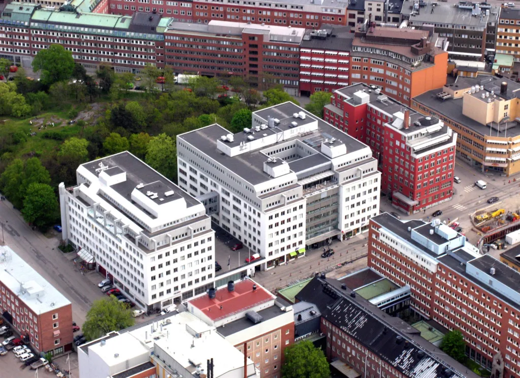 Flygbild över vita och röda byggnader omgivna av grönska och stadsstruktur.