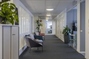 1. En kontorskorridor med fåtöljer, växter och glasväggar längs ena sidan.