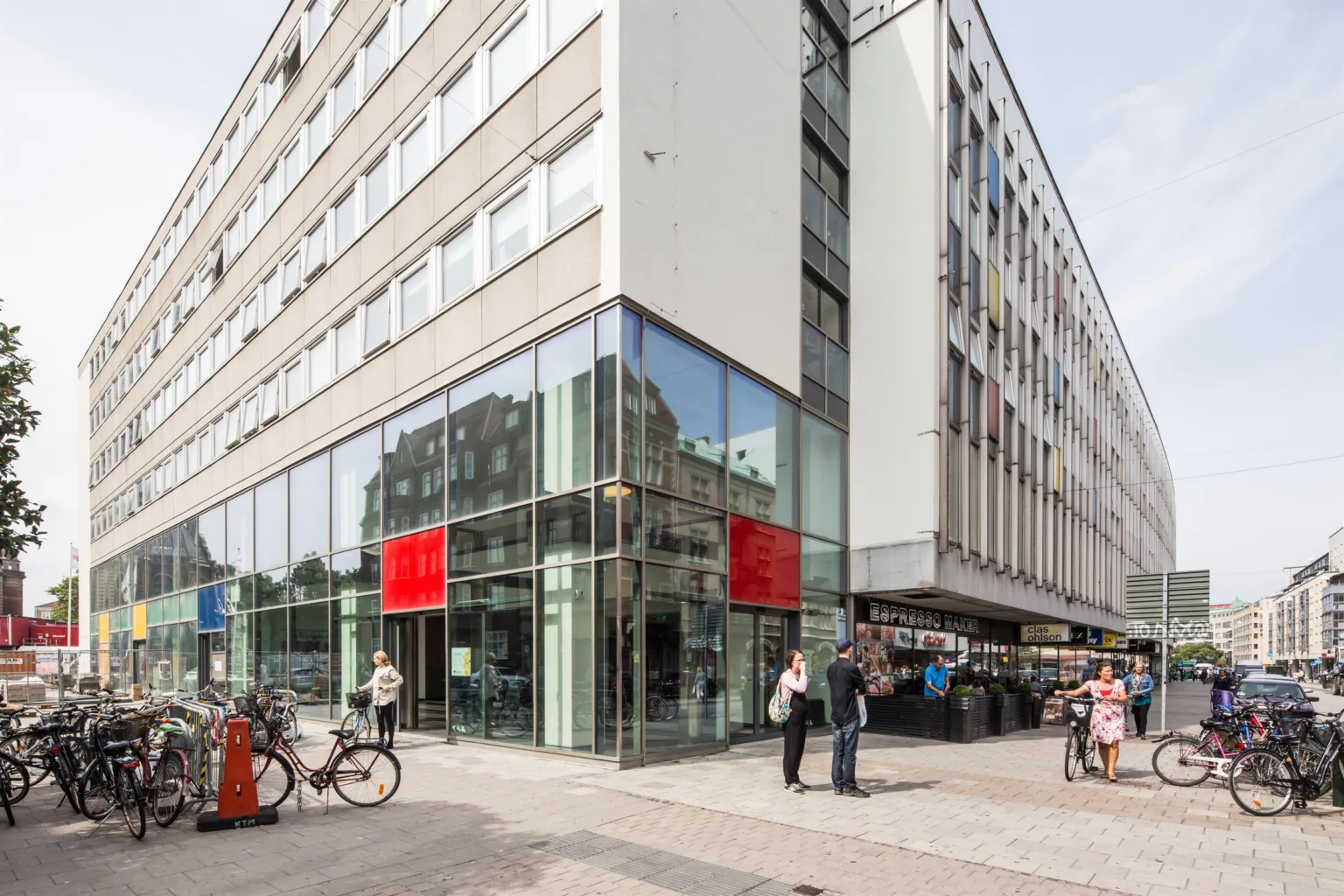 En hörnbyggnad med glasfasad och röda skyltar, människor och cyklar vid trottoaren.