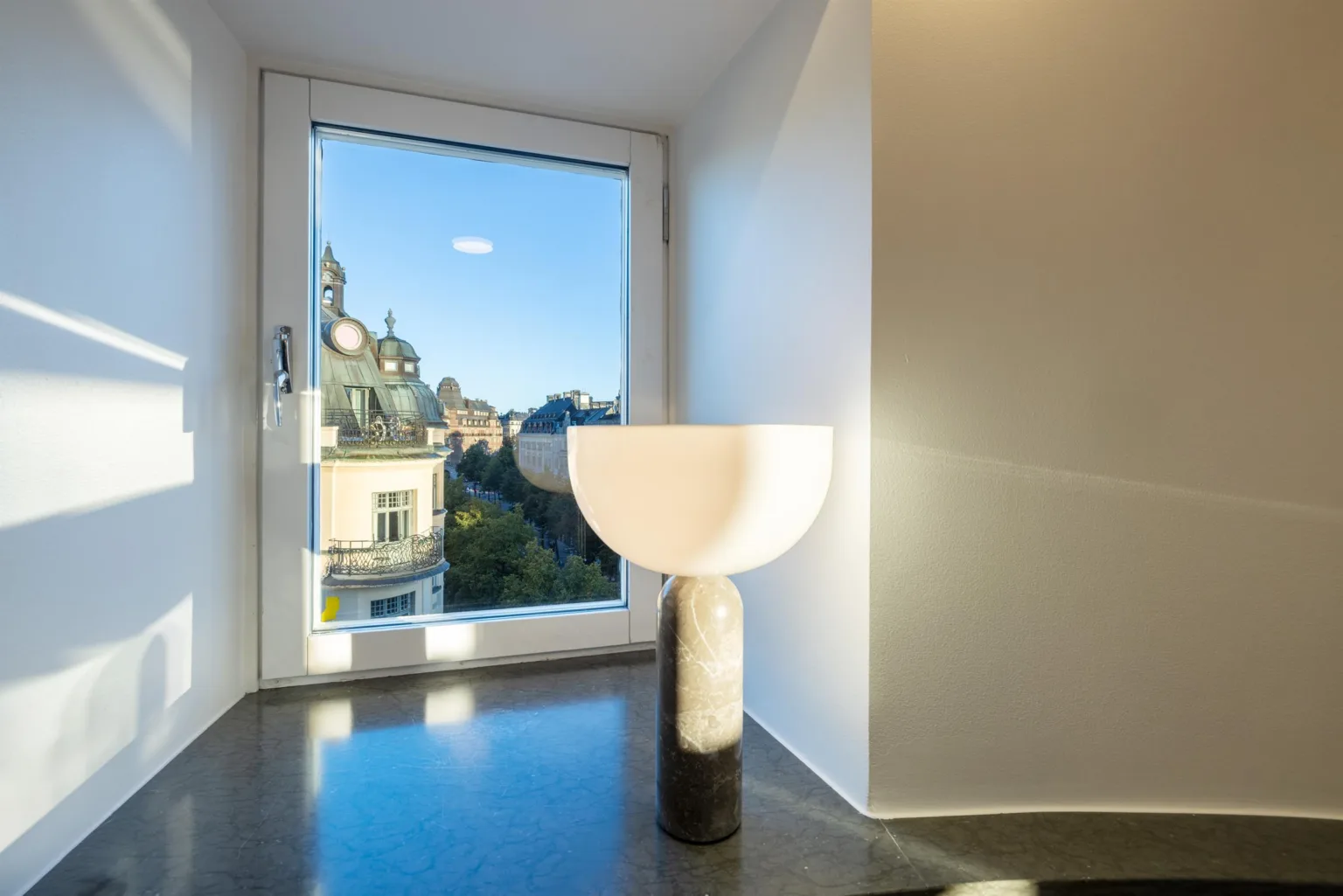 4. En vit bordslampa med rund skärm står på en mörk yta bredvid ett stort fönster med utsikt över en byggnad.