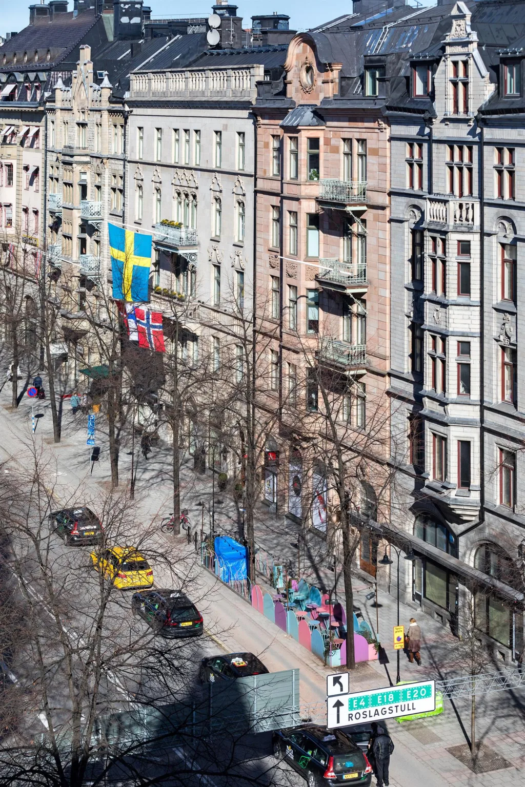 En stadsgata med klassiska byggnader, svenska flaggan på en fasad och bilar parkerade längs trottoaren. Träd utan löv kantar gatan.