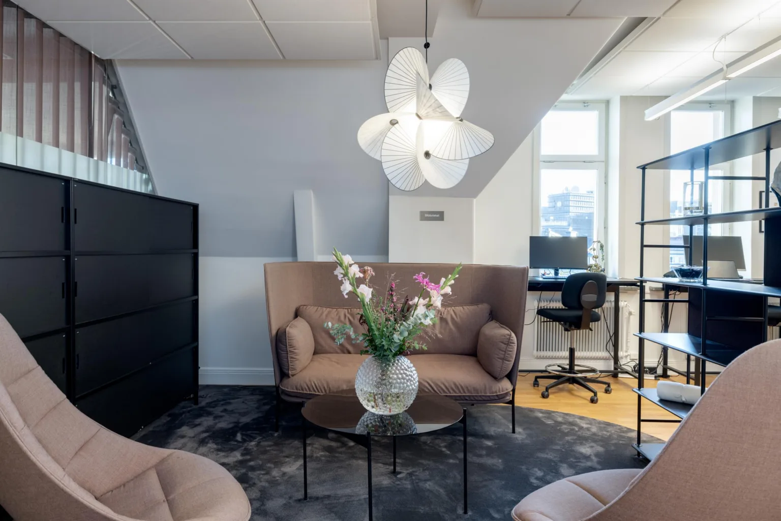 En mysig sittgrupp med beige soffa, blommor i vas på ett bord och en dekorativ lampa i taket.