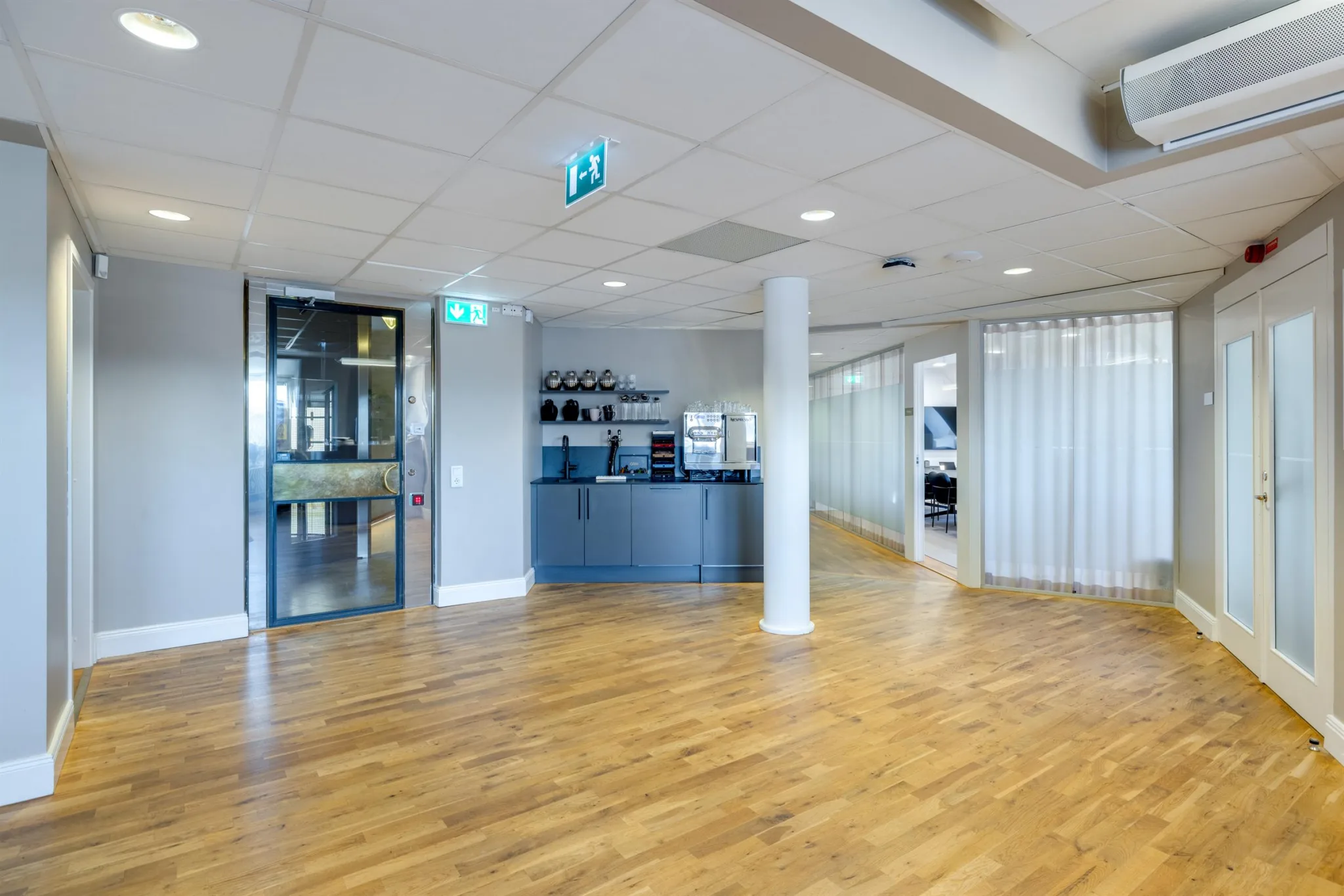 Ett öppet kontorsutrymme med trägolv, en receptionsdisk och glasdörrar i bakgrunden. Nödutgångsskyltar syns i taket.