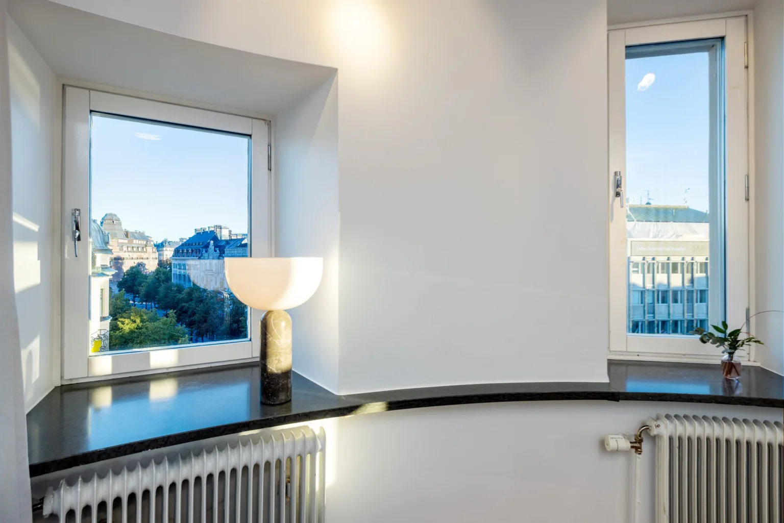 5. Ett hörnrum med rundad fönsterbänk, en bordslampa och två fönster med utsikt över stadsmiljö.