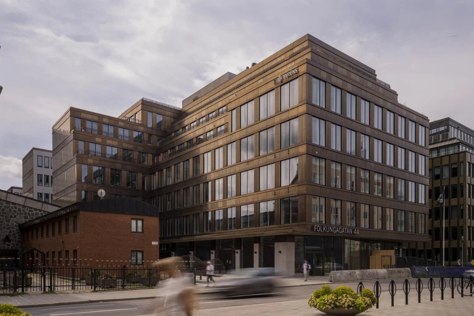 En tegelbyggnad med stora glasfönster och en mindre byggnad bredvid, samt människor och bilar på gatan.