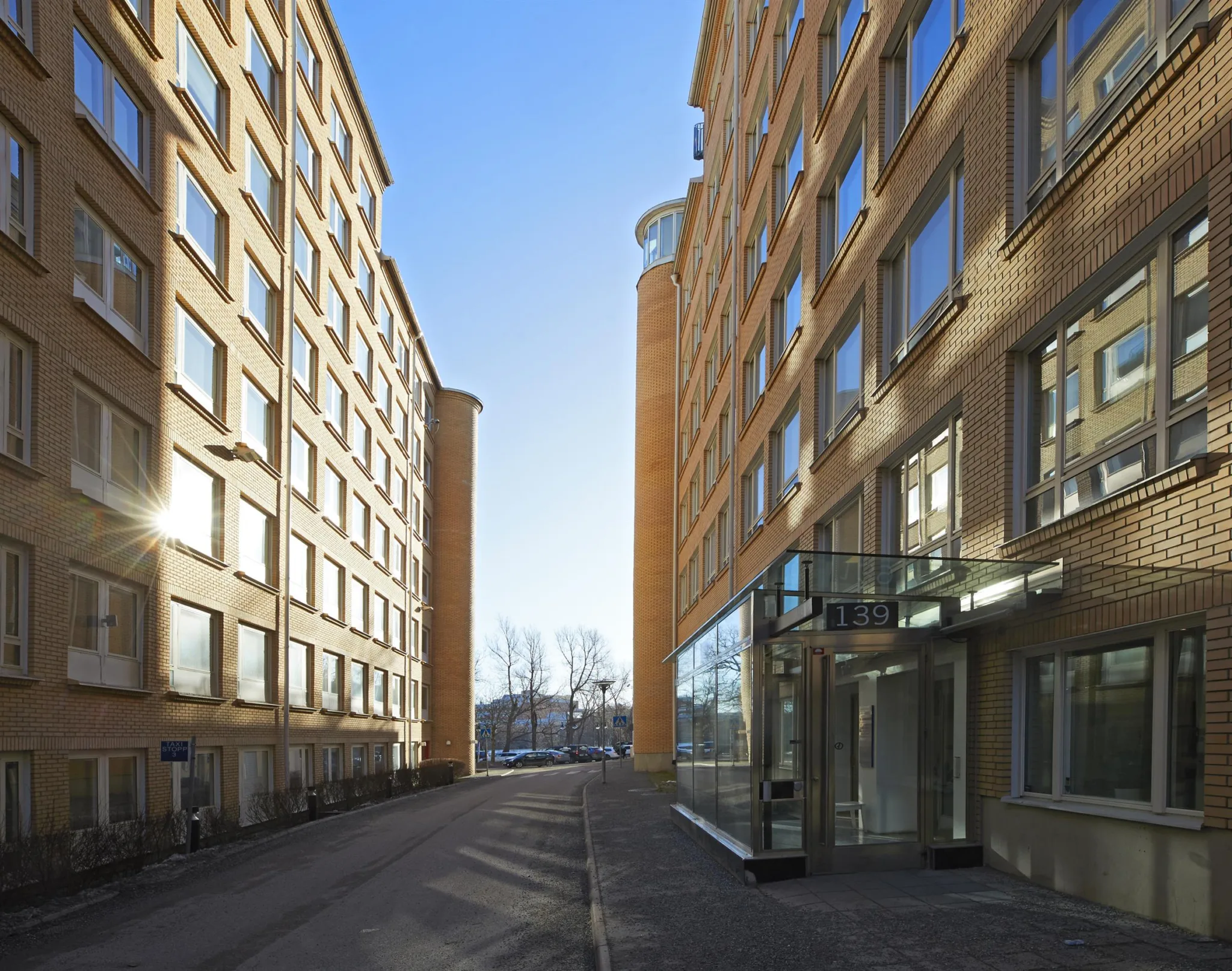4. Två höga tegelbyggnader med stora fönster, som bildar en smal gata i mitten, upplyst av solen i bakgrunden.