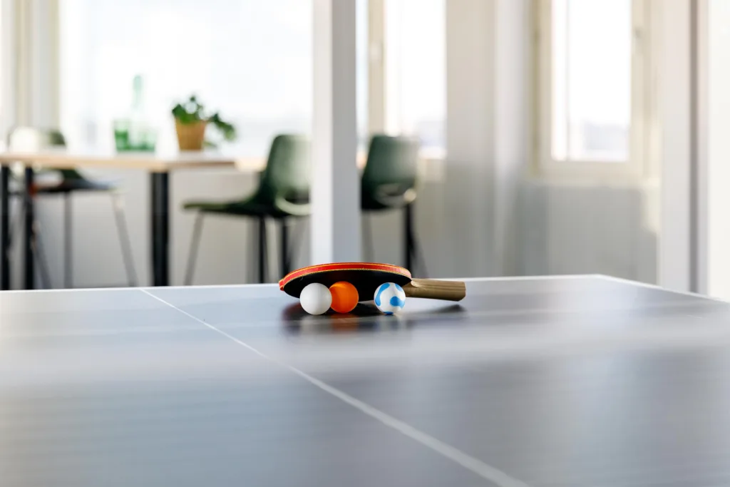 5. Ett pingisracket och tre bollar ligger på ett bord, med suddiga stolar och ett fönster i bakgrunden.