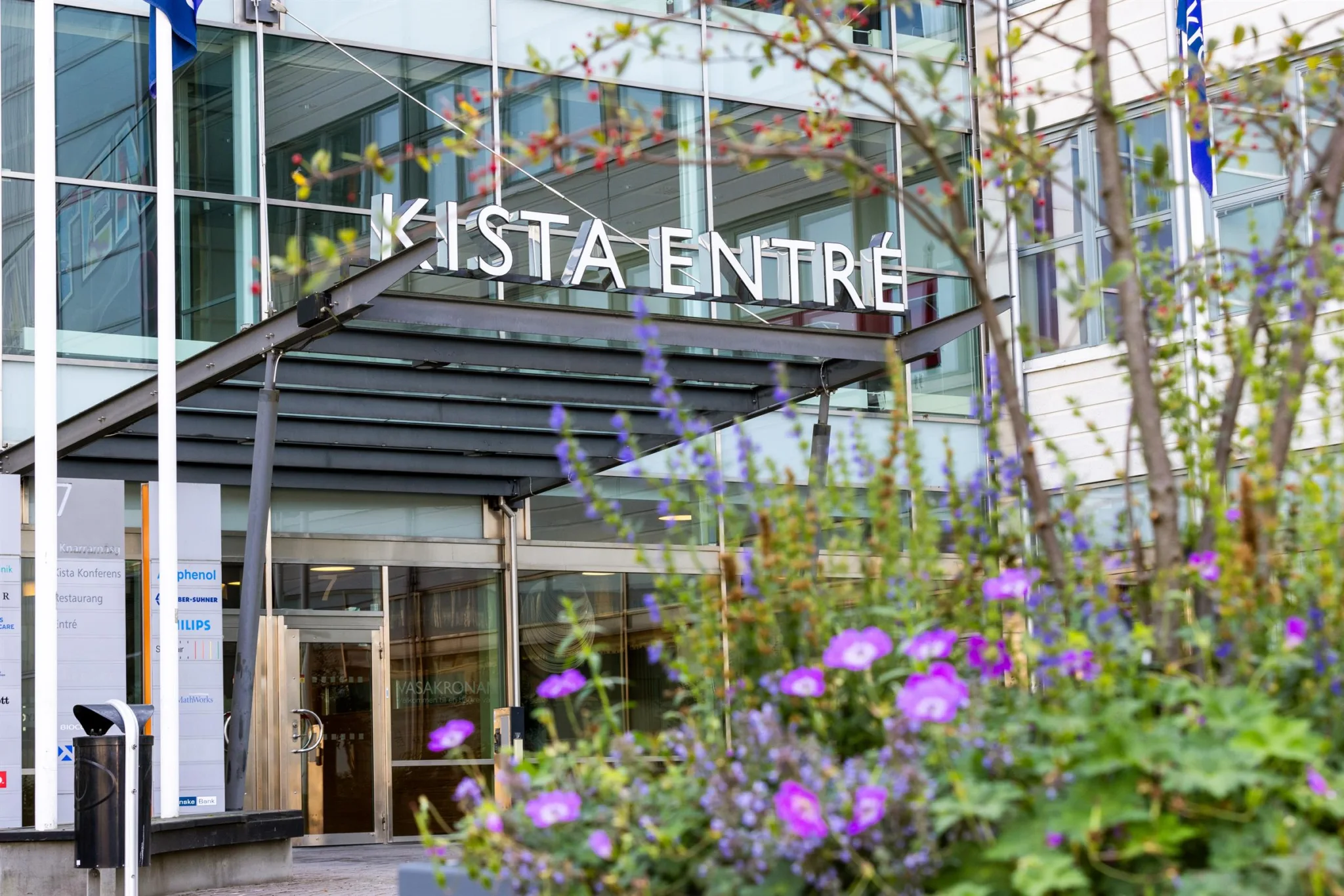 4. Entré till "Kista Entré" med en glasfasad, taköverhäng och lila blommor i förgrunden.