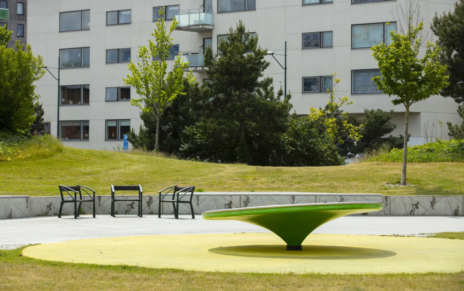 En grön lekstruktur i en park med bänkar och flerbostadshus i bakgrunden.