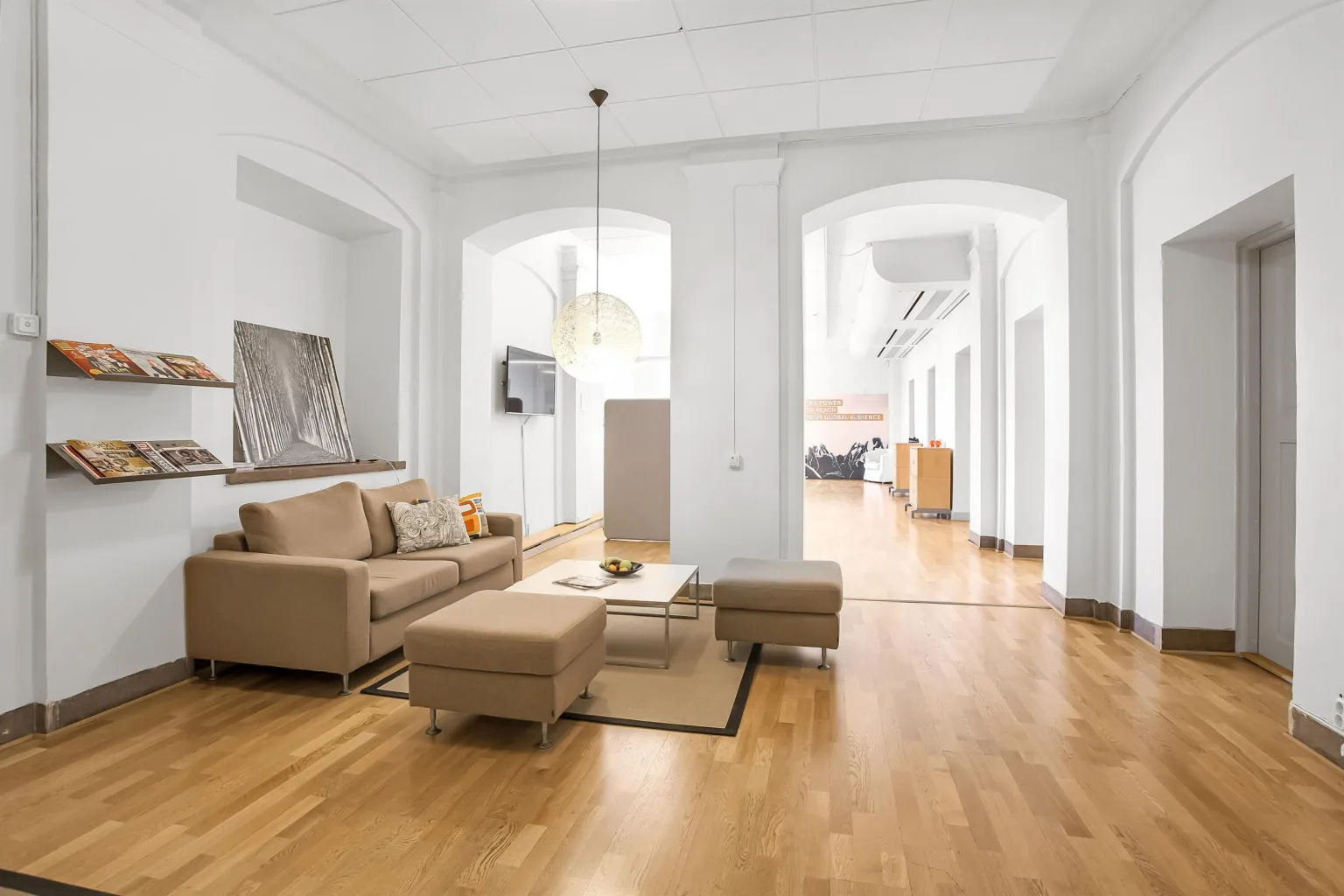 Ett ljust vardagsrum med beige soffa, soffbord och träparkettgolv. I bakgrunden syns en öppen planlösning med valvbågar.