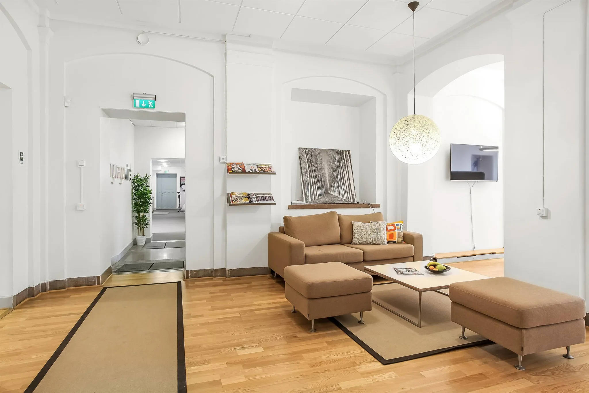 Ett modernt vardagsrum med beige soffa, soffbord och en hängande rund lampa. I bakgrunden syns en dörröppning och hyllor med tidningar.