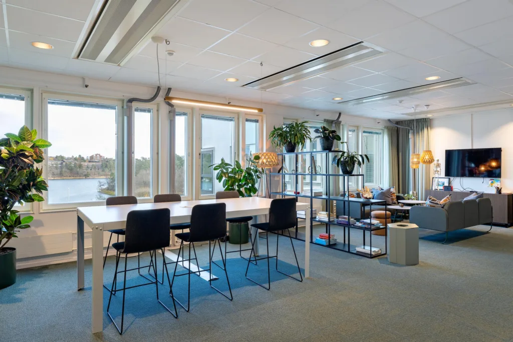 2. Ett gemensamt utrymme med ett högt bord och barstolar, grönväxter och hyllor mot stora fönster.