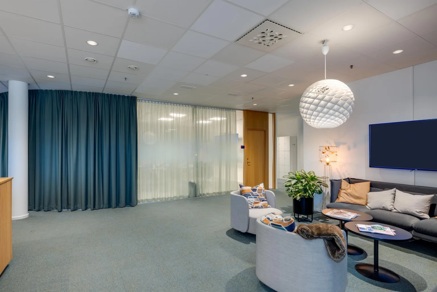 4. Ett loungområde med soffor, ett soffbord, en växt och modern belysning, intill blå gardiner och glasväggar.