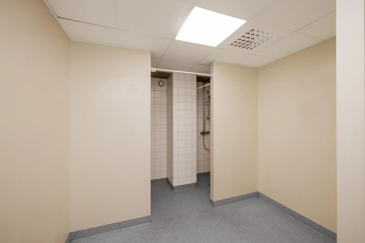 3. Ett litet rum med beige väggar, grått golv och en öppen garderobsdel med rör synliga.
