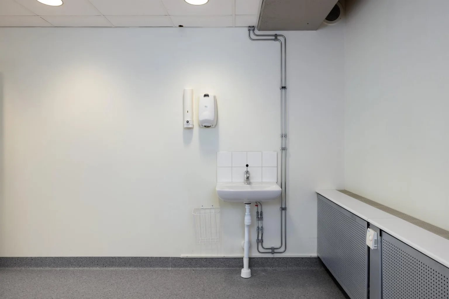 En vit handfat med kran på en vägg, omgiven av två vita väggmonterade dispensrar och synliga rör.