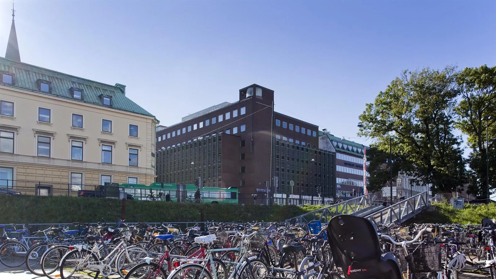 En stadsmiljö med flera cyklar i förgrunden och byggnader i olika stilar, inklusive en brun tegelbyggnad.
