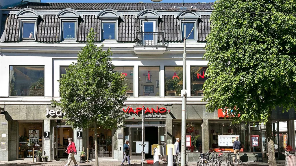 En byggnad med en butik på bottenvåningen, omgiven av träd och gångväg. Text på byggnaden: "KAPAND".