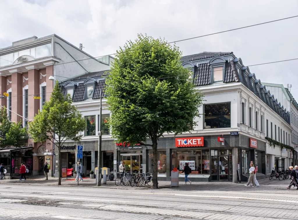En byggnad med skyltfönster och texten "TICKET", omgiven av väg och trottoar med cyklar och träd.