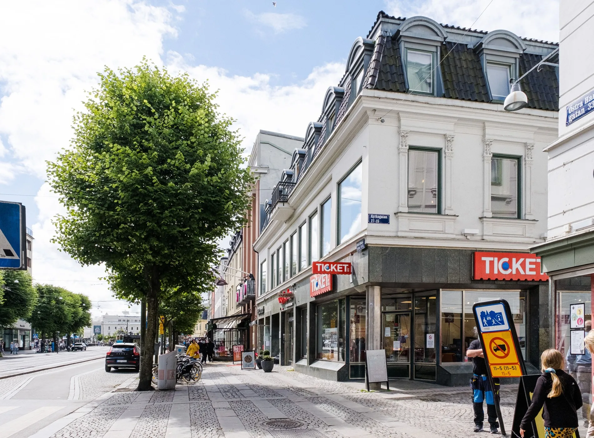 En stadsgata med äldre byggnader, butiker och en trottoar kantad av träd.
