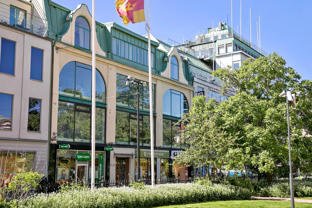 En fasad med flera butiksingångar, flaggor, stora glaspartier och träd i förgrunden.