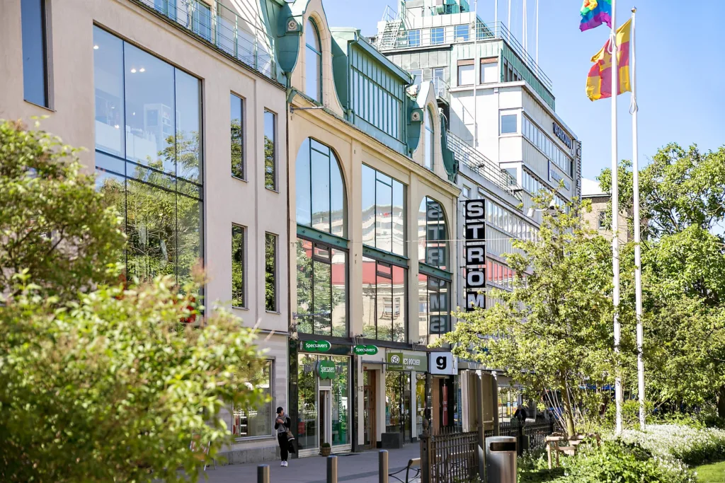 En affärsbyggnad med skyltar, flaggor, glasfasader och grönska runt omkring.