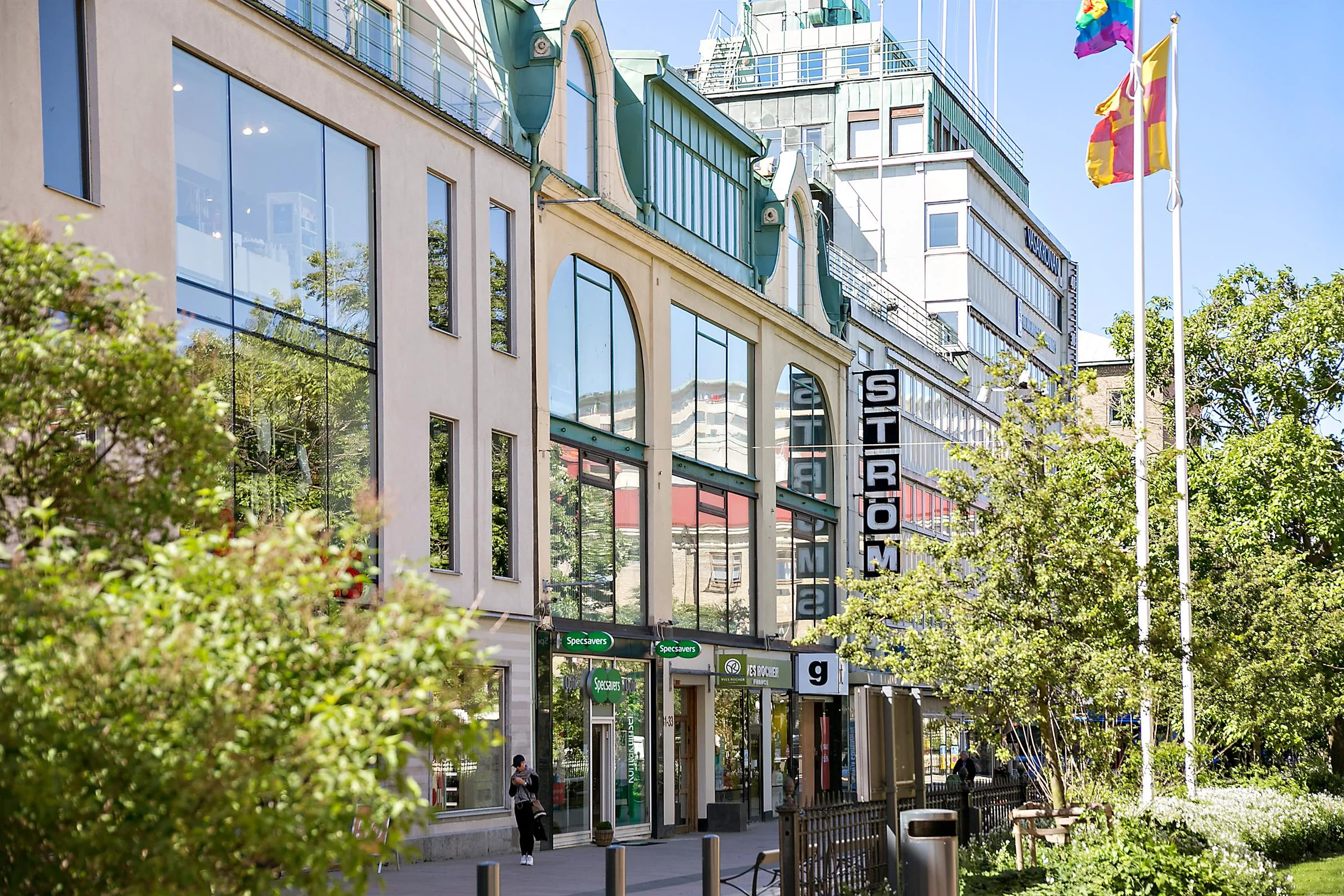 En affärsbyggnad med skyltar, flaggor, glasfasader och grönska runt omkring.
