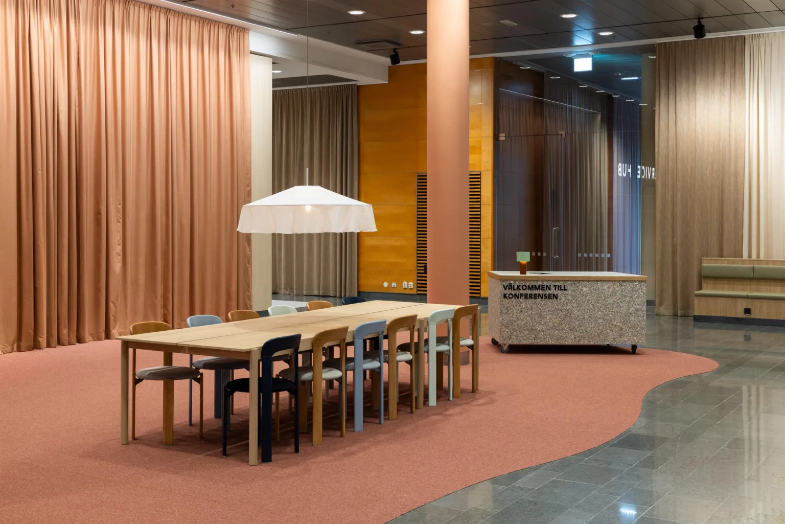 3. Ett modernt inomhusutrymme med ett långt bord, stolar och en vit taklampa, omgiven av rosa gardiner och en reception.