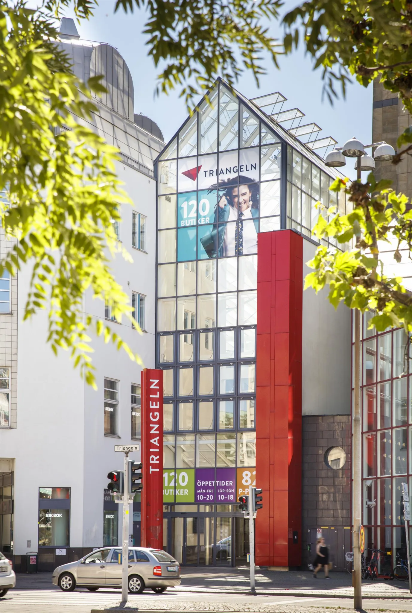 1. En modern byggnad med glasfasad och röda detaljer, omgiven av träd och andra byggnader.